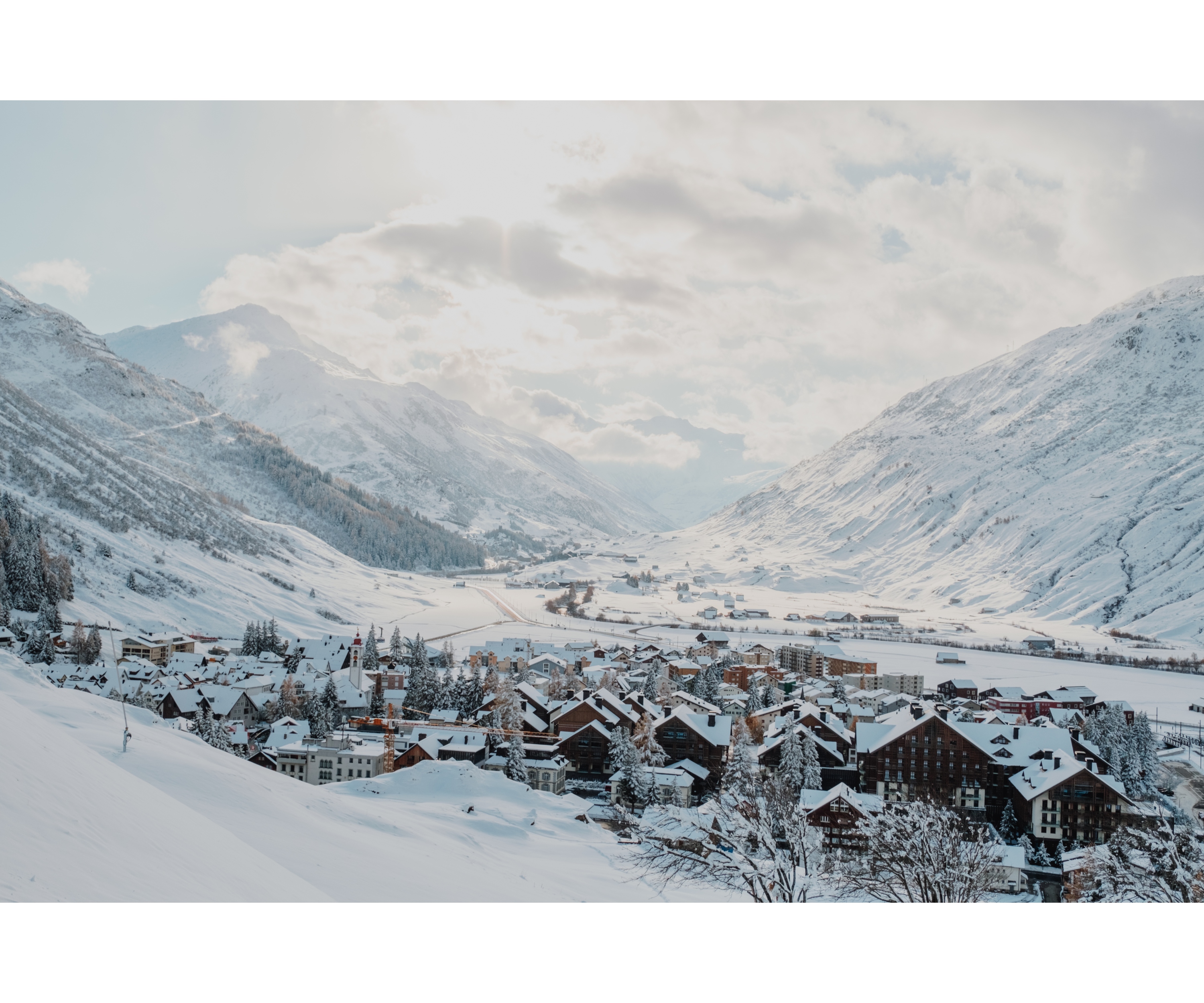 Photo over Andermatt at sunrise. The sky is slightly cloudy and the mountains in the background and the whole landscape is covered with snow.