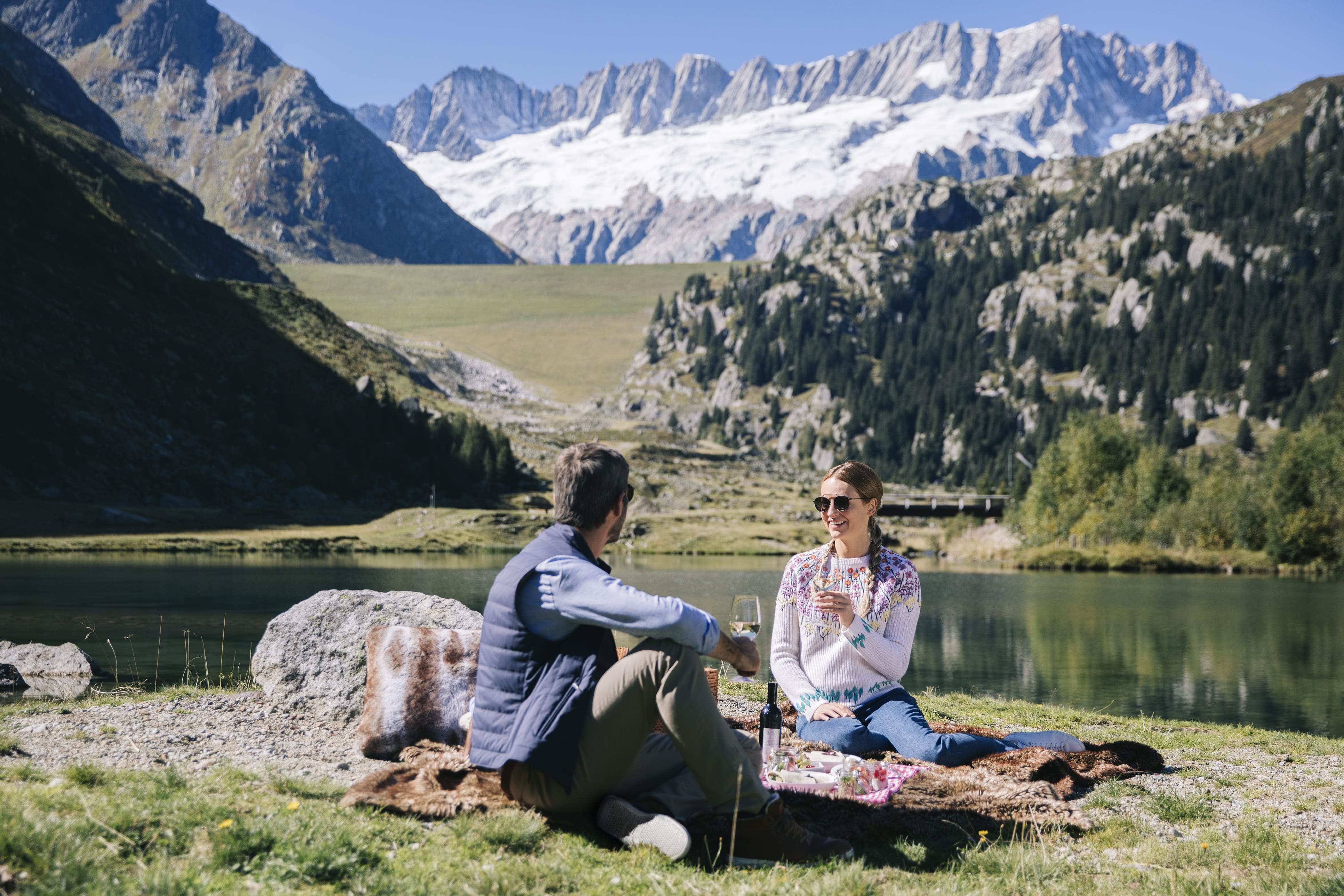 A Man And Woman Sitting On A Rock By A Lake With Mountains In The Background